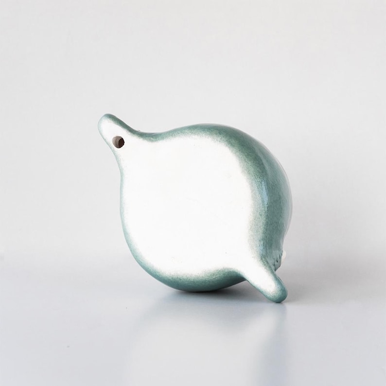 May include: A ceramic, teardrop-shaped decorative object with a hole at the top and a small protrusion at the bottom. The object has a glossy, two-tone finish, with a white center and a teal-green edge.
