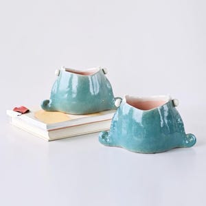 May include: Two turquoise ceramic planters with a unique shape, featuring small white accents and a pink interior. The planters have a textured surface and are placed on a white surface next to a stack of books.