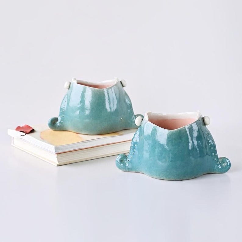 May include: Two turquoise ceramic planters with a unique shape, featuring small white accents and a pink interior. The planters have a textured surface and are placed on a white surface next to a stack of books.