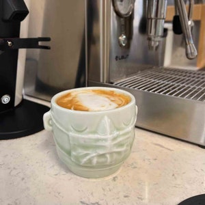 May include: A green ceramic mug with a textured design resembling a face, filled with a light brown coffee drink with a layer of white foam on top.