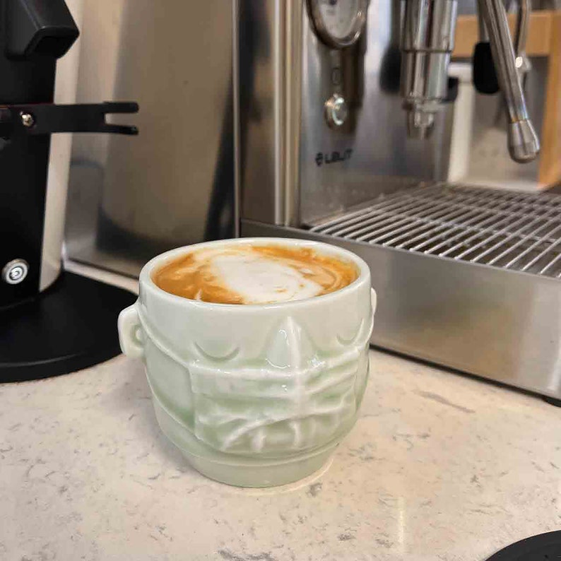 May include: A green ceramic mug with a textured design resembling a face, filled with a light brown coffee drink with a layer of white foam on top.