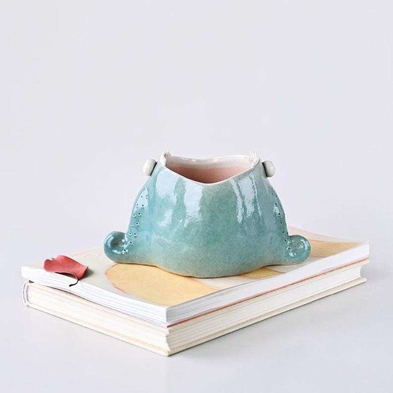 May include: A turquoise ceramic vessel with a unique shape, featuring small, raised dots and two rounded protrusions. The interior is a soft pink. It rests on a stack of books with a red leaf. Decorative pottery.