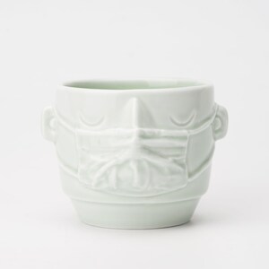 May include: A white ceramic mug with a face and a mask sculpted on the front.