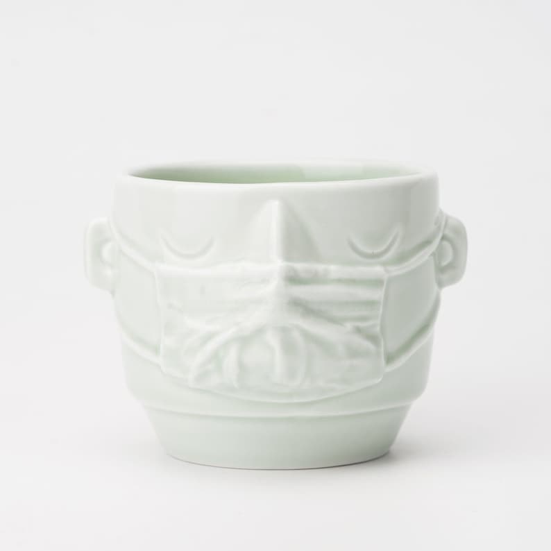 May include: A white ceramic mug with a face and a mask sculpted on the front.