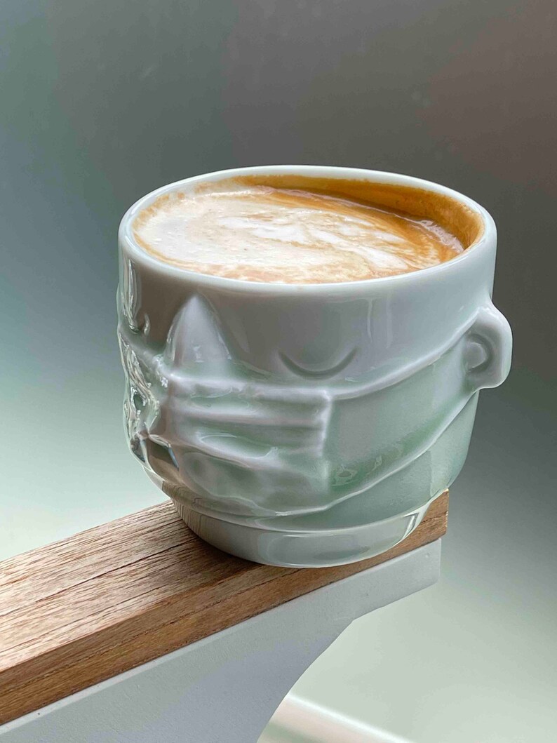 May include: A light green ceramic coffee mug with a face design. The mug has a white rim and is filled with coffee.