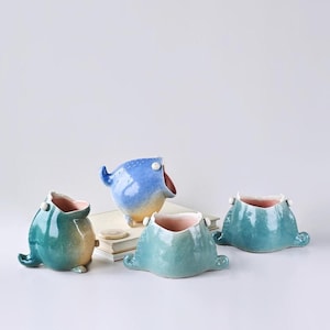 May include: Four whimsical ceramic planters in the shape of open-mouthed creatures. The planters are glazed in shades of blue and green, with pink interiors. One is blue with yellow feet, and another has a tail. They sit on a white surface.