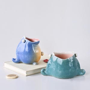 May include: Two ceramic planters in the shape of whimsical creatures. One is blue with a yellow belly, the other is teal. Both have pink interiors and small, rounded feet. They sit on a book and a white surface.