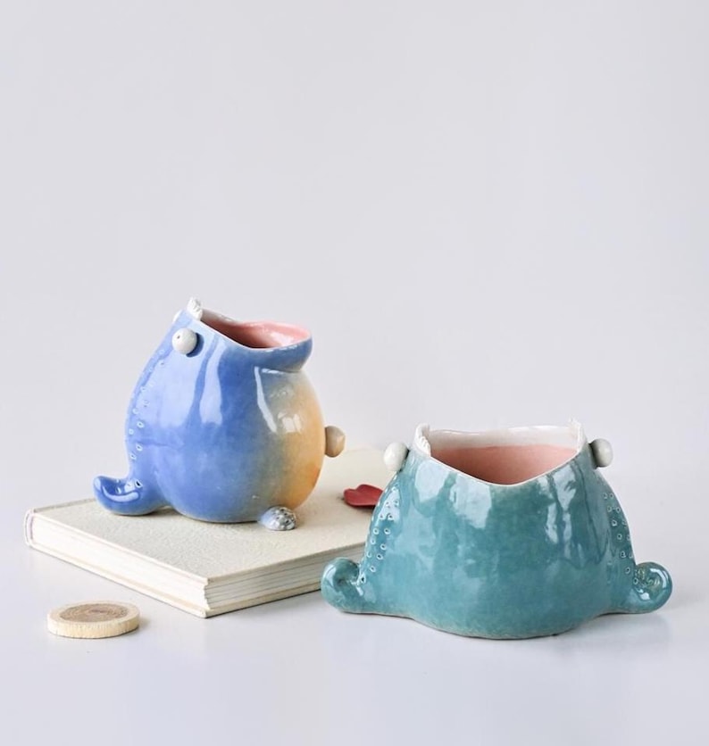 May include: Two ceramic planters in the shape of whimsical creatures. One is blue with a yellow belly, the other is teal. Both have pink interiors and small, rounded feet. They sit on a book and a white surface.