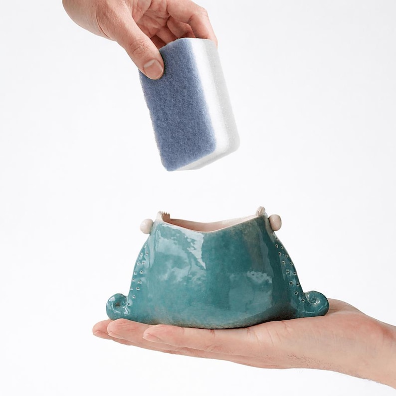 May include: A teal ceramic vessel with a unique shape, held in a hand, with a blue and white sponge being lowered into it. The vessel has a textured surface and decorative elements. The background is plain white.