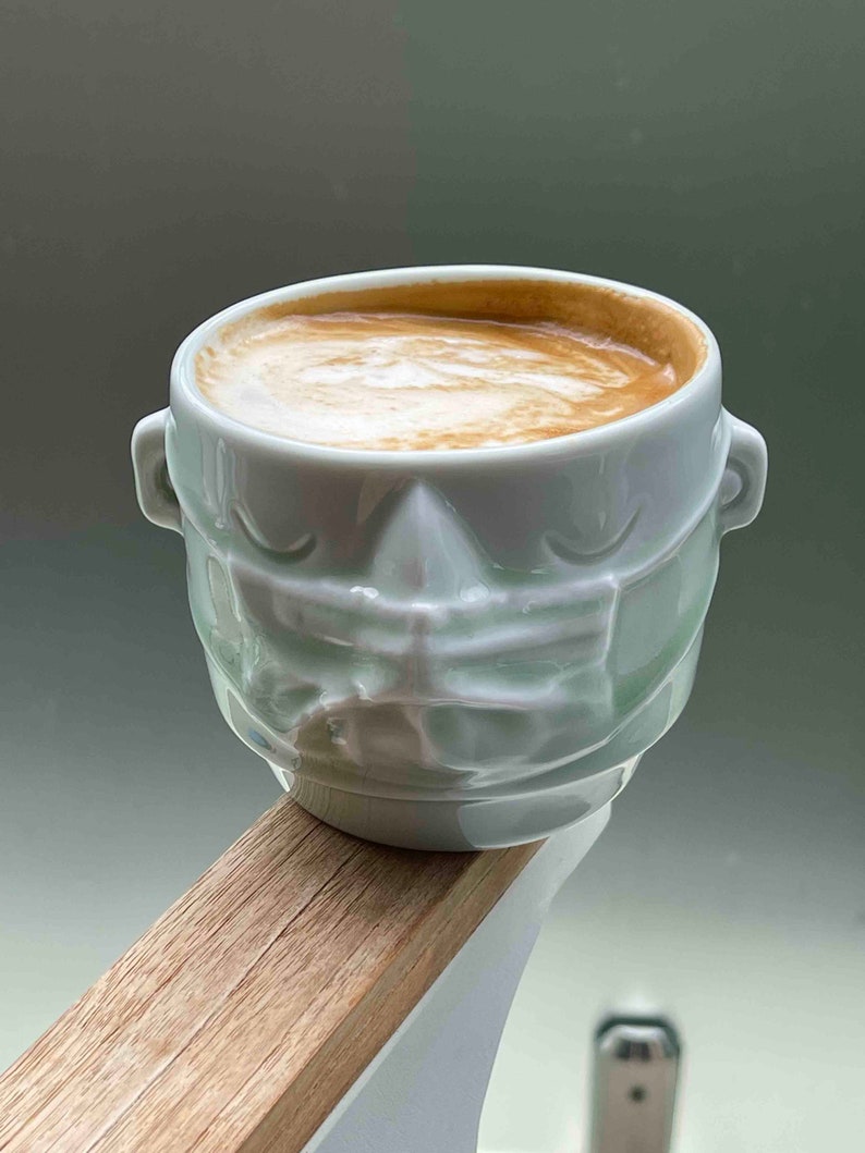 May include: A light green ceramic coffee mug with a textured face design. The mug is filled with coffee and has a layer of foam on top.