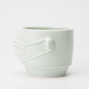 May include: A light green ceramic mug with a raised design of a hand covering the mouth. The mug is a unique and quirky piece of pottery.