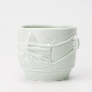 May include: A light green ceramic mug with a face and a white face mask sculpted on the front.