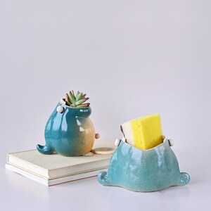 May include: Two ceramic whale-shaped containers. One teal and yellow container holds a succulent plant. The other teal container holds a yellow and white sponge. Both containers have small, white, round accents.