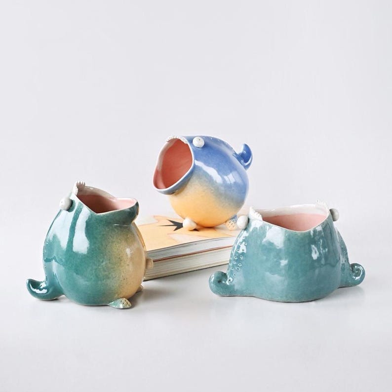 May include: Three whimsical ceramic planters in the shape of stylized creatures. The planters are glazed in shades of blue, teal, and yellow, with pink interiors. Each has a wide opening and small, rounded feet. One is perched on a book.