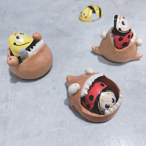 Peut inclure: Quatre figurines en céramique à la bouche ouverte, chacune tenant un chocolat différent. Les bonbons sont en forme d'abeille, de coccinelle et de deux coccinelles. Les figurines sont peintes en brun, blanc et noir.