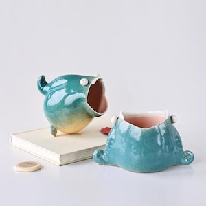 May include: Two whimsical ceramic planters. One is teal with an open mouth and small legs. The other is a teal, squat design with a pink interior. Both have a glossy finish and are on a white surface.