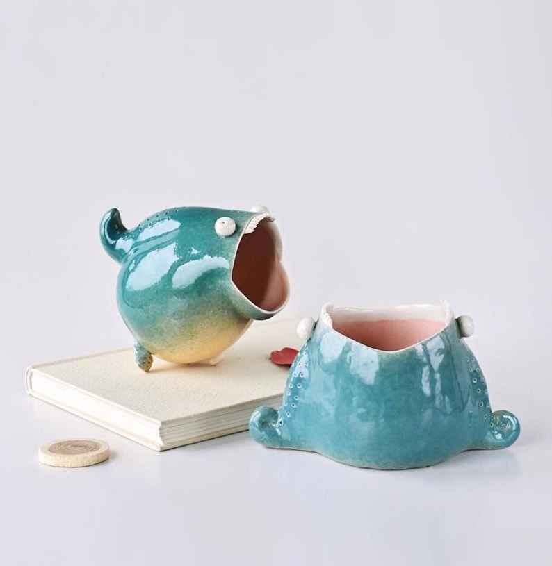 May include: Two whimsical ceramic planters. One is teal with an open mouth and small legs. The other is a teal, squat design with a pink interior. Both have a glossy finish and are on a white surface.