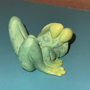 May include: A green and yellow ceramic frog figurine with a textured surface. The frog is curled up in a sleeping position.