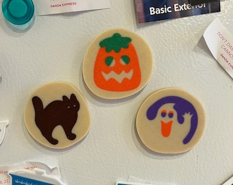 May include: Three Halloween-themed magnets on a white refrigerator. One magnet features a brown cat silhouette, another an orange jack-o'-lantern, and the third a purple ghost with an orange mouth. The magnets are round and beige.