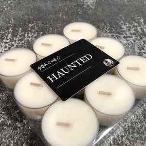 Puede incluir: Un envase de plástico transparente que contiene ocho velas de té blancas con mechas de madera. Una tarjeta negra con la palabra "HAUNTED" en letras blancas está colocada encima. También es visible la marca "Apotheka Candle Co.".