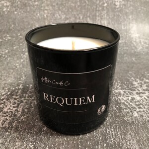Puede incluir: Una vela de vidrio negro con un interior de cera blanca y una mecha de madera. La vela tiene una etiqueta que dice "Requiem" y "Apotheka Candle Co."