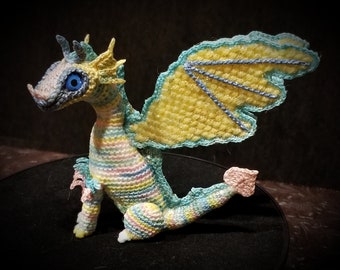 Könnte beinhalten: Ein gehäkeltes Drachenspielzeug mit einem gelb, blau und rosa gestreiften Körper und einem gelben und blauen Flügel. Der Drache hat ein blaues Auge und einen rosa Schwanz.