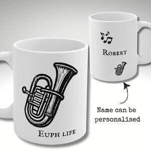 Puede incluir: Dos tazas de cerámica blanca. Una presenta una ilustración en blanco y negro de un eufonio con el texto "Euph Life". La otra taza muestra notas musicales, el nombre "Robert" y una pequeña ilustración de eufonio. El texto dice "Name can be personalised".