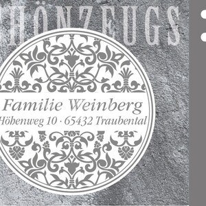 Op de afbeelding: Een ronde, witte rubberen stempel met een grijs bloemenmotief en de tekst "Familie Weinberg" en "Höhenweg 10. 65432 Traubental".