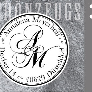 Pode incluir: Um carimbo redondo, branco, auto-entintador com borda preta. O carimbo apresenta um monograma preto das letras "A" e "M" em uma fonte de letra cursiva decorativa. O texto "Annalena Meyerhoff" e "Dorfstr. 14, 40629 Düsseldorf" é impresso em torno do monograma em uma fonte sans-serif preta.