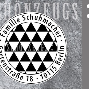 May include: A round stamp featuring a black and white triangle design. The stamp is inscribed with the text "Familie Schuhmacher" and "Gartenstraße 18 - 10115 Berlin". It has a wooden handle.
