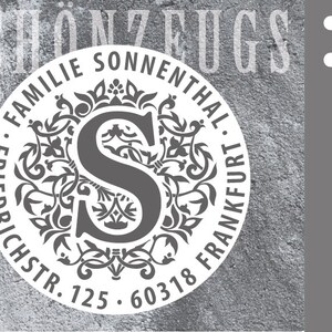 Könnte beinhalten: Ein runder weißer Stempel mit einem schwarzen floralen Design und dem Buchstaben "S" in der Mitte. Der Text um das Design lautet "Familie Sonnenthal, Friedrichstr. 125, 60318 Frankfurt."