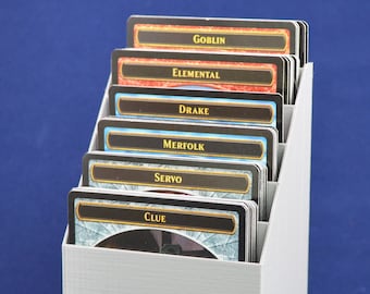 May include: A gray plastic card holder with six slots for cards. The cards are labeled with the words "Goblin", "Elemental", "Drake", "Merfolk", "Servo", and "Clue".