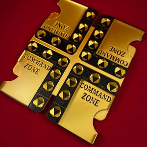 May include: A gold-colored game board with four sections, each labeled "Command Zone". Each section has a grid of black and gold hexagonal spaces with text labels like "Life", "CMDR", and "DMG".