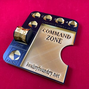 May include: A gold-colored plastic command zone game tray with a black border. The tray has a slot for a dice tower and a space for a dice. The tray also has a space for cards and a space for tokens. The tray is labeled "Command Zone" and "Realityfoundry.net".