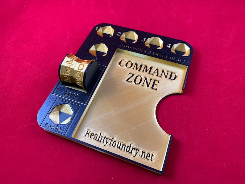 May include: A gold-colored plastic command zone game tray with a black border. The tray has a slot for a dice tower and a space for a dice. The tray also has a space for cards and a space for tokens. The tray is labeled "Command Zone" and "Realityfoundry.net".