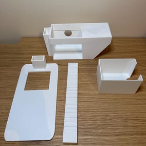 May include: A white 3D printed model of a house with a separate roof, stairs, and a small rectangular structure. The model is assembled on a wooden surface.