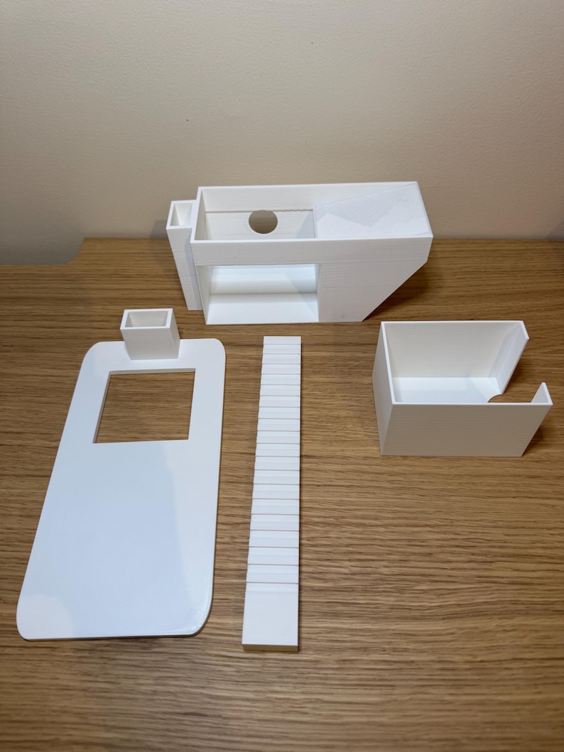 May include: A white 3D printed model of a house with a separate roof, stairs, and a small rectangular structure. The model is assembled on a wooden surface.