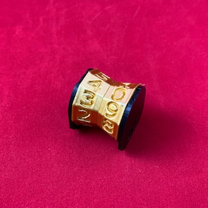 May include: A gold-colored, multi-sided, numbered dice with black edges. The dice is made of plastic and has the numbers 2, 3, 4, 5, 6, 8, 9, and 0 printed on its sides.