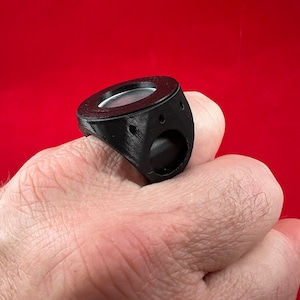 May include: A black 3D printed ring with a round, silver-coloured insert. The ring is worn on a person's finger.