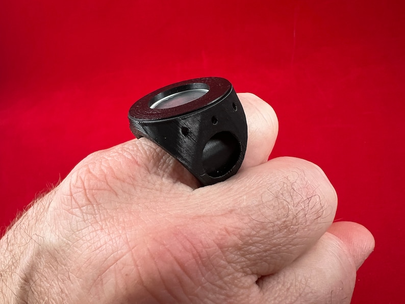 May include: A black 3D printed ring with a round, silver-coloured insert. The ring is worn on a person's finger.