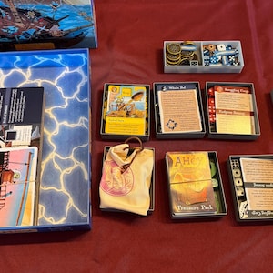 May include: A board game with various components, including cards, dice, tokens, and a game board. The game box features a ship illustration. The game pieces are arranged on a red surface.