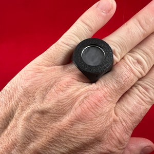 May include: A black ring with a round, textured, black face and a small, clear window in the centre.