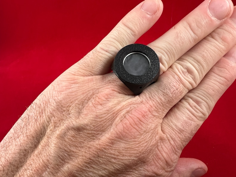 May include: A black ring with a round, textured, black face and a small, clear window in the centre.