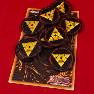 Könnte beinhalten: Ein Stapel von acht schwarzen und goldenen Spielsteinen mit einem dreieckigen Design und einem kleinen Kreis in der Mitte. Die Spielsteine liegen auf einer Yu-Gi-Oh! Trading Card Game Karte.