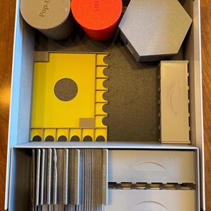May include: A gray box with a foam insert for organizing board game components. The insert has compartments for cards, tiles, and other game pieces. The box also includes two round containers labeled "Hills" and "Pop-Ups".
