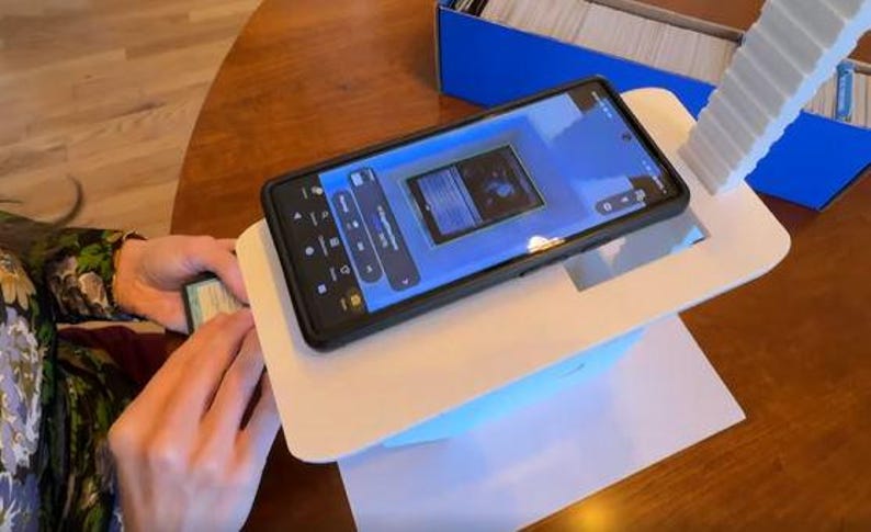 May include: A white phone stand with a phone on top of it. The phone is displaying a card game app. The stand is on a wooden table.