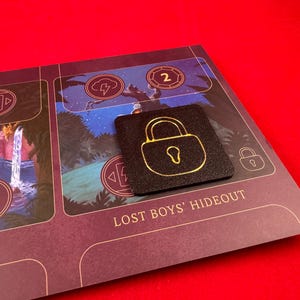 May include: A black square game piece with a gold padlock design. The piece is on a purple board game with the text "Lost Boys' Hideout".