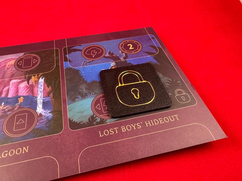 May include: A black square game piece with a gold padlock design. The piece is on a purple board game with the text "Lost Boys' Hideout".