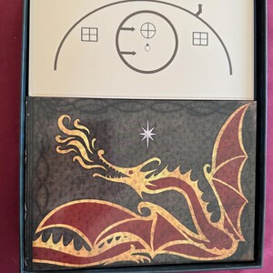 May include: A board game box with a black frame. The top card features a house illustration with a circular door and two windows. The game box below has a dragon illustration in gold and red.