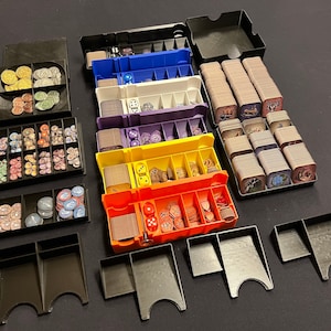 May include: A set of black plastic storage boxes with dividers for organising game pieces. The boxes contain various coloured tokens, dice, and cards.